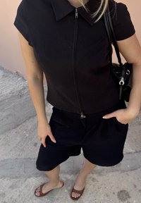 Mulher com camisa preta com fecho e calções, a levar uma mala preta ao ombro, em cima de um passeio de betão, com sandálias castanhas tipo slide.