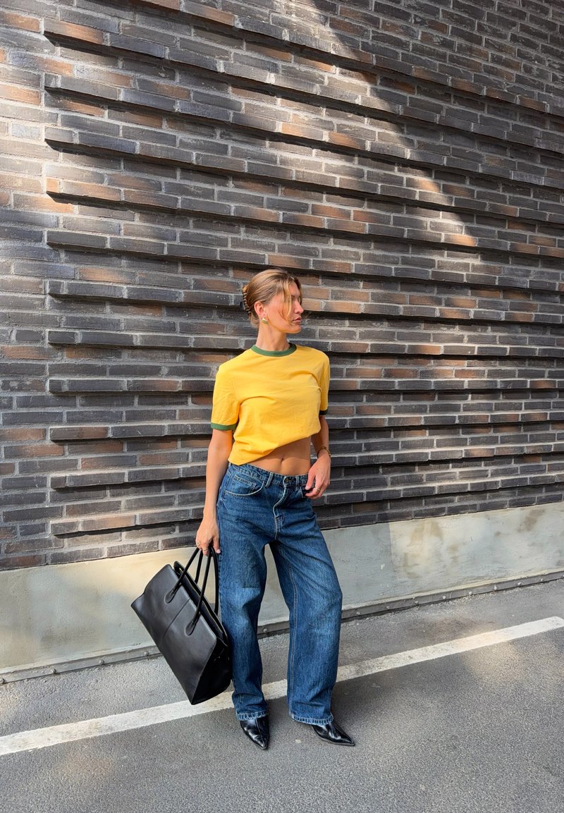 Femme en crop top jaune, jean large bleu et chaussures noires pointues, porte un grand sac à main noir, debout contre un mur de briques texturé.