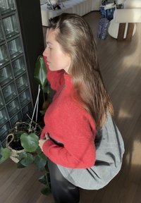 Egy piros, texturált pulóver, egy szürke táska és sötét nadrág, mellettük egy zöld növény, a háttérben egy üvegtégla fal.