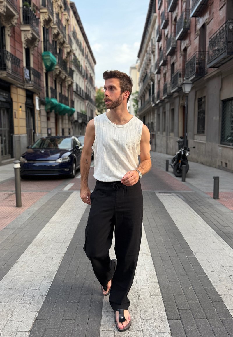 Jeune homme en pull sans manches blanc et pantalon noir marchant dans une rue urbaine avec une voiture et une moto garées en arrière-plan.