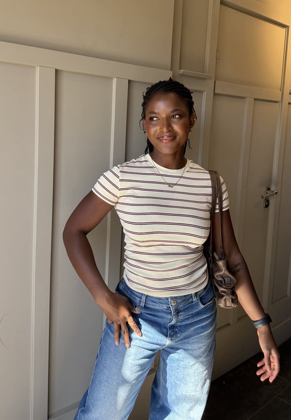 Mujer joven con el cabello trenzado que lleva una camiseta blanca a rayas, jeans azules y un bolso de hombro, de pie en el interior con la mano en la cadera.