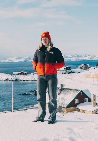 Chaqueta de plumón negra y roja con cremallera, jeans grises y un gorro naranja brillante, usados al aire libre en un paisaje costero nevado.