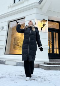 Svart boblejakke med hette trimmet med fuskepels, vertikal kanaldesign, parret med et rutete skjerf og strikket lue, plassert i et snødekt miljø.