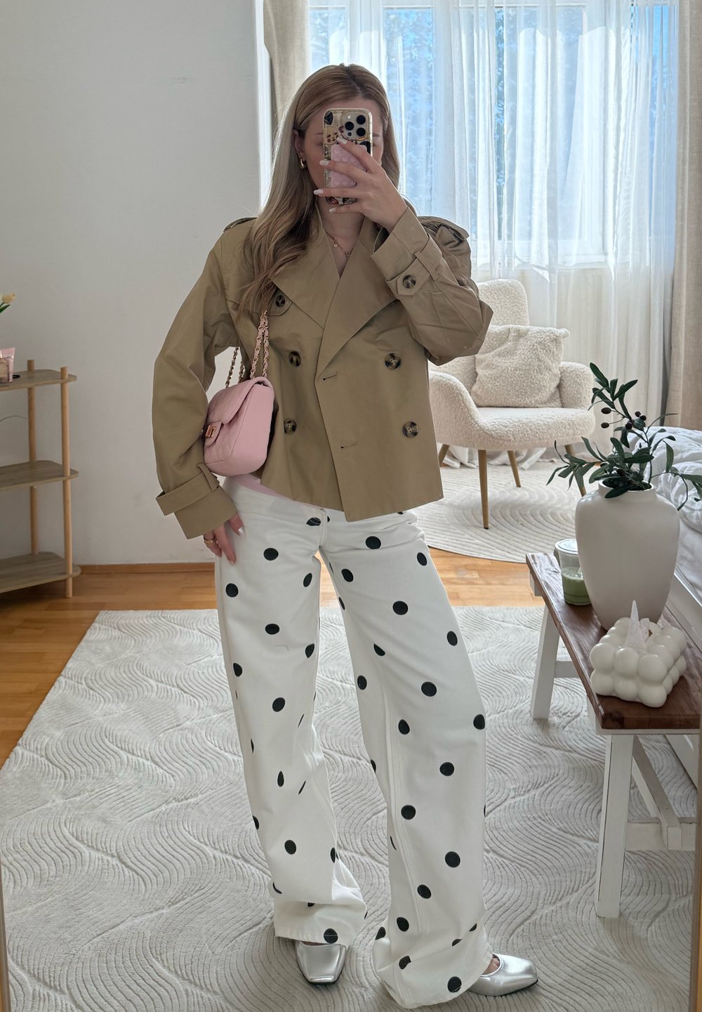 Femme portant une veste croisée beige, un pantalon large blanc à pois noirs, des chaussures argentées, tenant un sac rose dans un salon lumineux.