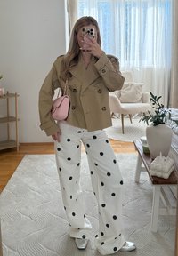 Femme portant une veste croisée beige, un pantalon large blanc à pois noirs, des chaussures argentées, tenant un sac rose dans un salon lumineux.