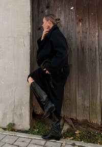 Zwart overjas, jurk tot de knie in zwart met een split, en zwarte laarzen tot boven de knie van leer. Gladde textuur met subtiele ontwerpelementen.