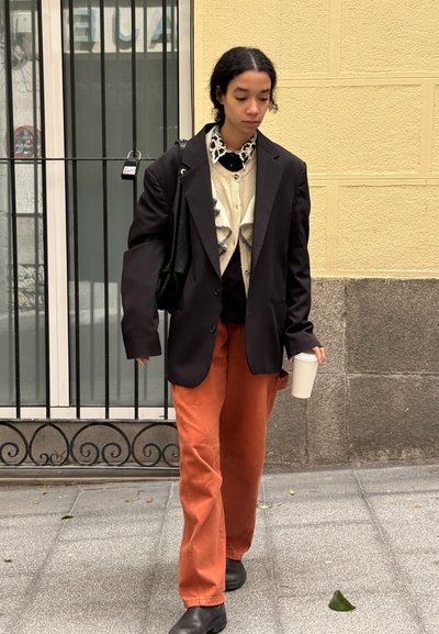 Blazer negro oversize, cárdigan crema con volantes, camisa blanca, pantalones naranjas de pierna ancha y botas negras. Sosteniendo una taza de café blanca.