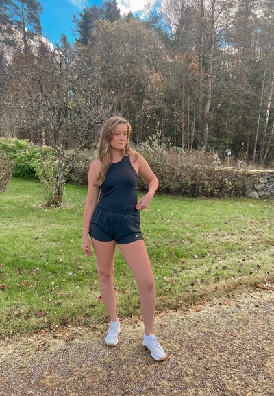 Mujer joven vestida con ropa deportiva negra, de pie sobre un camino de grava junto a un césped con árboles en un fondo otoñal.