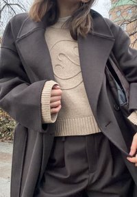 Ciemny wełniany płaszcz z szerokimi klapami nałożony na beżowy sweter dziany z fakturą. Brązowe spodnie z wysokim stanem dopełniają strój.