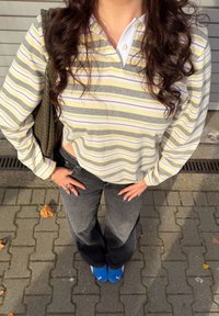 Osoba nosiaca žlté a sivé pruhované tričko s dlhým rukávom, čierne rifle, modré topánky a nesie na ramene zelenú tkanú tašku.