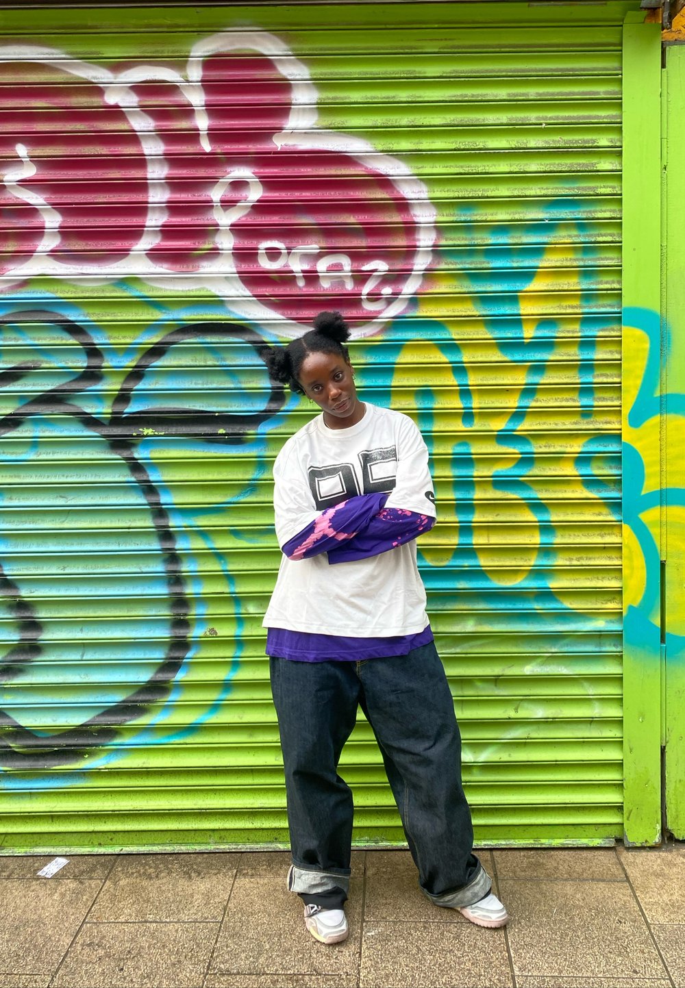 T-shirt branca com estampa preta, usada por cima de uma camisa de manga comprida roxa, calças de ganga largas e sapatilhas cinzentas. Fundo de graffiti brilhante.