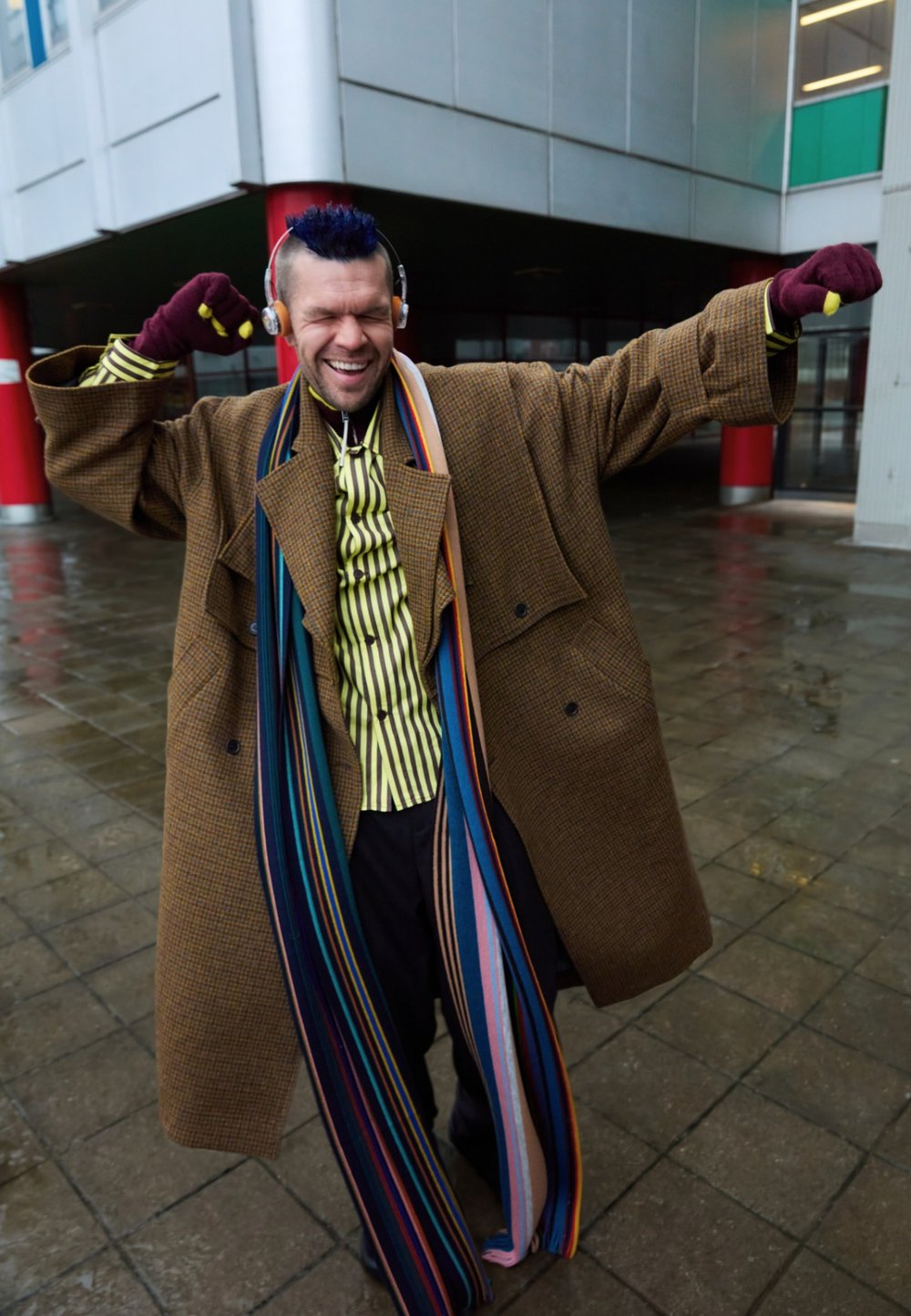 Gabardina marrón de pata de gallo, bufanda larga de rayas multicolor, camisa abotonada de rayas verticales, auriculares y guantes.