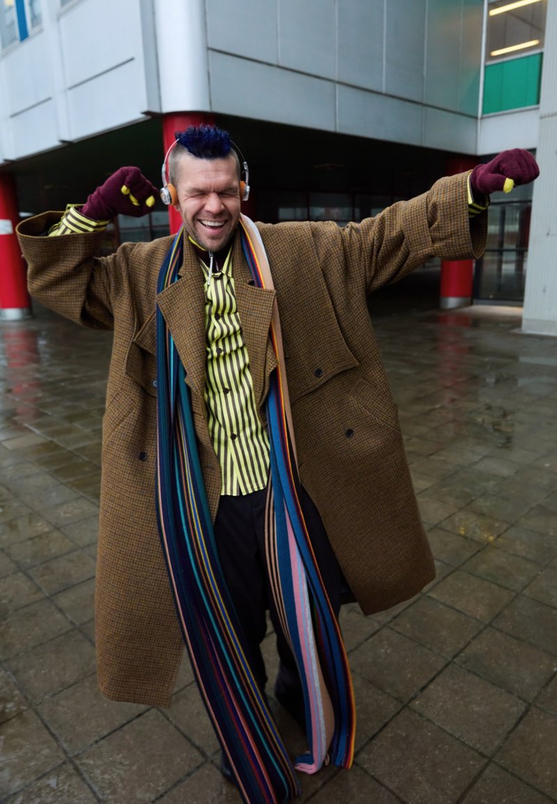 Manteau à chevrons marron, longue écharpe rayée multicolore, chemise à boutons à rayures verticales, casque audio et gants.