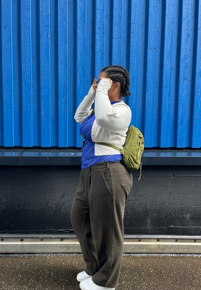 Mochila verde, camisa de rayas azul, cárdigan blanco y pantalones de pierna ancha oscuros. Cabello trenzado, de pie contra una pared azul acanalada.