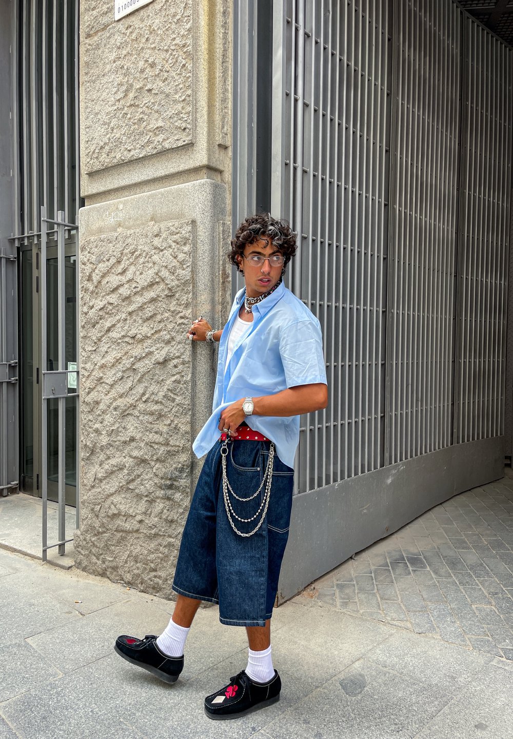 Tânăr cu păr creț purtând o cămașă albastră, pantaloni scurți lungi din denim cu lanțuri, șosete albe și pantofi negri mergând lângă o poartă metalică.