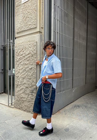 Joven con cabello rizado vistiendo una camisa azul, pantalones cortos de mezclilla largos con cadenas, calcetines blancos y zapatos negros caminando cerca de una puerta de metal.