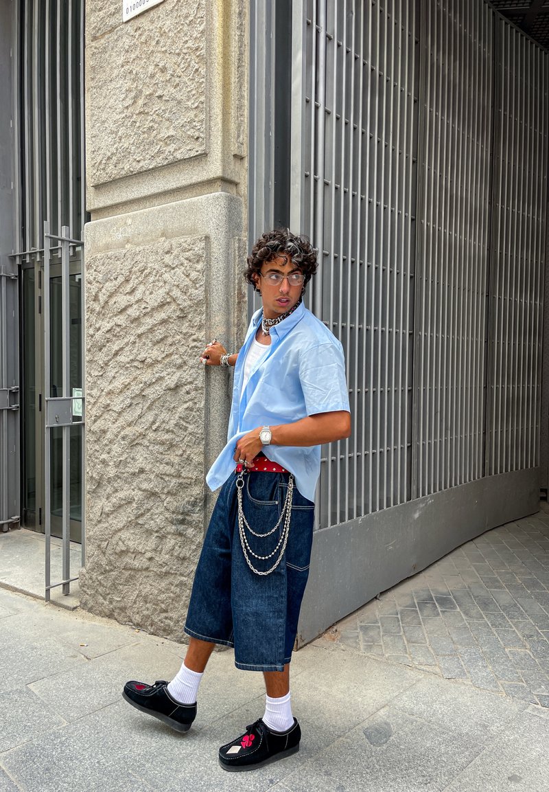 Young man with curly hair wearing blue shirt, long denim shorts with chains, white socks, and black shoes walking near metal gate.