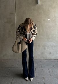 Ζακέτα με τιάφρο λουλούδια, λευκή μπλούζα, φαρδές σκούρο τζιν παντελόνι και γκρι τσάντα. Τα μυτερά ψηλοτάκουνα παπούτσια ολοκληρώνουν το σύνολο. Σοβαρός φόντος από μπετό.