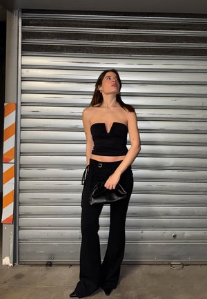 Mujer con top negro sin tirantes y pantalones de pierna ancha, de pie sosteniendo un clutch negro, con una persiana metálica cerrada detrás de ella.