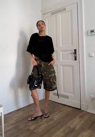 Mujer vistiendo una blusa negra transparente y pantalones cortos de camuflaje, sosteniendo un bolso negro, de pie sobre un suelo de madera delante de una puerta blanca.