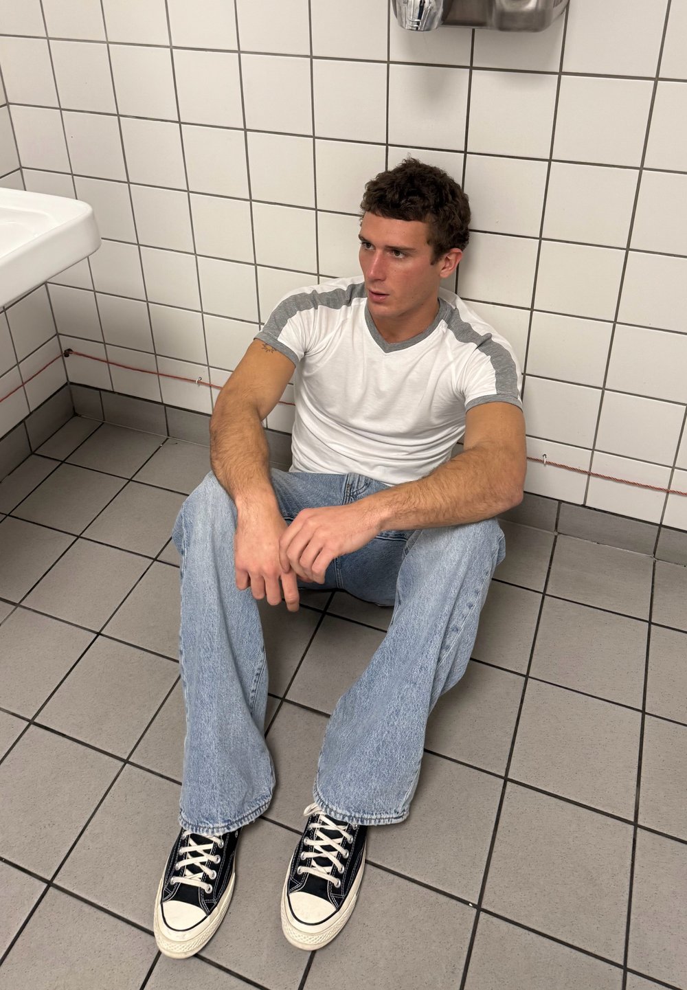 Νέος άντρας με λευκό και γκρι μπλουζάκι και τζιν κάθισε στο πλακάκι του μπάνιου, στηρίζεται στον πλακιδωτό τοίχο και κοιτάζει προς το πλάι.