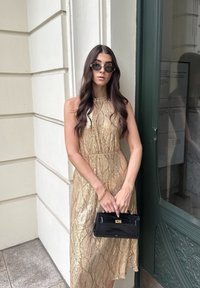 Mujer con pelo largo y oscuro, lleva gafas de sol y un vestido sin mangas dorado con lentejuelas, sostiene un bolso negro y está de pie junto a una pared clara y una puerta verde.