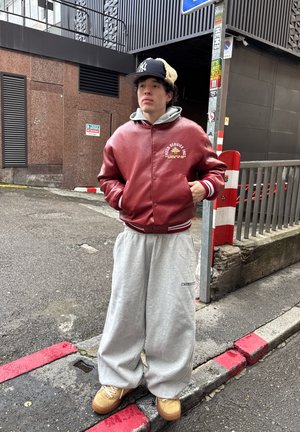 Joven de pie en una esquina de la calle vistiendo una chaqueta de cuero roja, pantalones deportivos grises, zapatos beige y una gorra negra al aire libre.