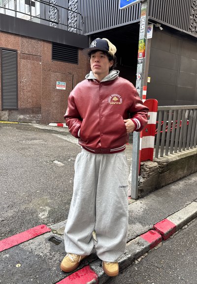 Joven de pie en una esquina de la calle vistiendo una chaqueta de cuero roja, pantalones deportivos grises, zapatos beige y una gorra negra al aire libre.