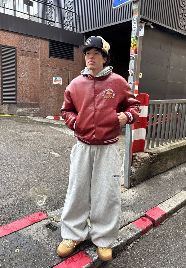 Jong persoon staand op een straatcorners met een rode leren jas, grijze joggingbroek, beige schoenen en een zwarte pet buiten.