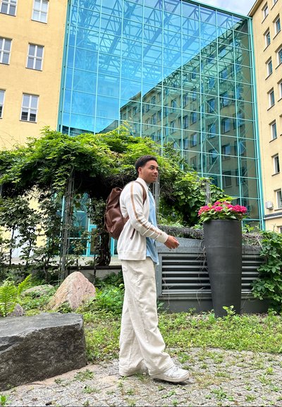 Una persona con un cárdigan gris claro sobre una camisa azul y pantalones crema está de pie cerca de una piedra, rodeada de vegetación y un jardinera de flores.