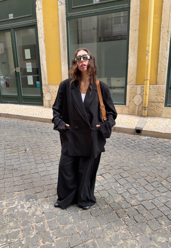 Una mujer con gafas de sol y un traje negro está de pie en una calle empedrada frente a unas tiendas amarillas, con un bolso marrón al hombro.
