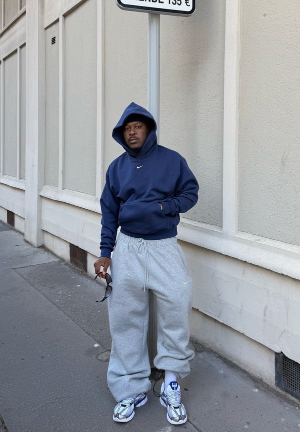Hombre con sudadera con capucha azul marino y pantalones deportivos grises, de pie en una acera de la ciudad sosteniendo gafas de sol, junto a una pared y una señal de tráfico.