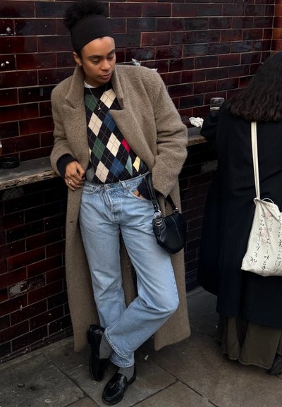 Persona apoyada contra una pared de azulejos oscuros, vistiendo un abrigo beige, suéter de rombos, jeans azul claro, mocasines negros y una diadema negra, con un pequeño bolso negro.