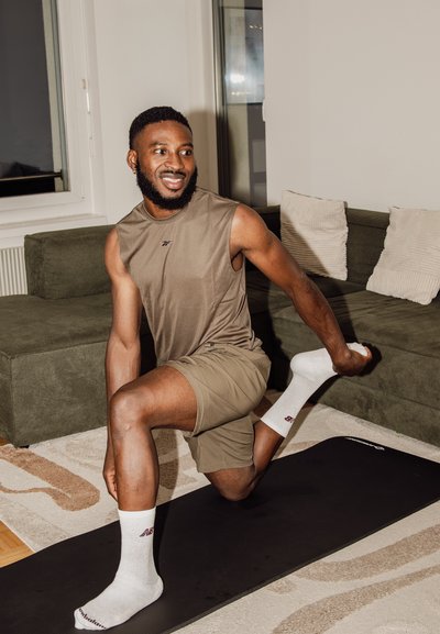 Hombre en ropa deportiva estirando las piernas en una esterilla de yoga en una sala de estar con un sofá verde y cojines blancos.