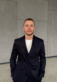 Fiatal férfi rövid hajjal és szakállal, sötét kétsoros zakóban és fehér ingben, aki szürke csempézett fal előtt áll.