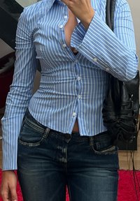 Ανοιχτό μπλε και άσπρη ριγέ πουκάμισο με μακριά μανίκια, εφαρμοστό σχέδιο και βαμβακερό ύφασμα, σε συνδυασμό με σκούρα τζιν παντελόνια.