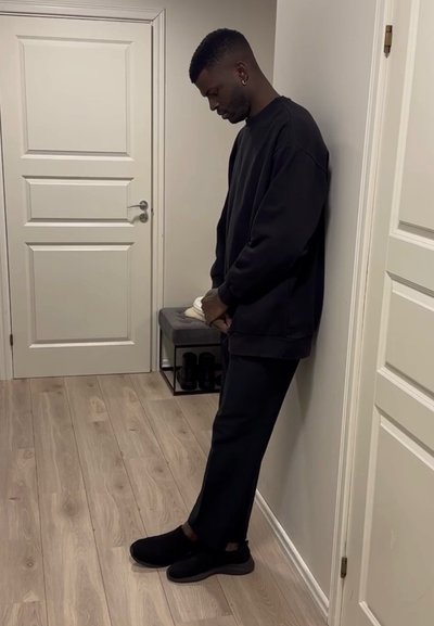 Hombre con atuendo negro apoyado contra la pared en un pasillo, sosteniendo zapatillas blancas junto a un pequeño banco con zapatos debajo.