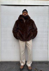 Giacca con cappuccio in pelliccia marrone dal tessuto spesso e morbido. Abbinata a pantaloni a quadri color beige chiaro e scarpe con plateau marroni.