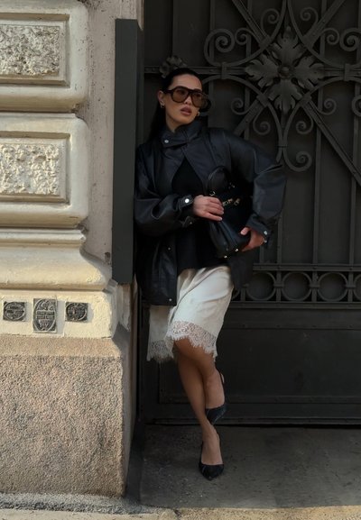 Mujer con gafas de sol grandes, chaqueta de cuero, falda blanca con encaje y tacones negros apoyada contra una puerta de metal ornamentada y una pared de piedra.