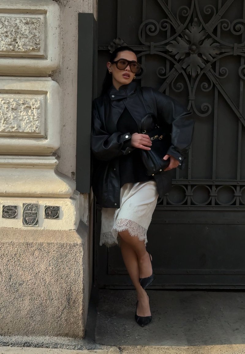 Woman wearing oversized sunglasses, leather jacket, white lace-trimmed skirt, and black heels leaning against ornate metal door and stone wall.