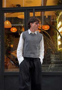 Pull sans manches gris tricoté porté sur une chemise blanche, associé à un pantalon large noir. L'arrière-plan présente un éclairage suspendu chaleureux et des plantes.