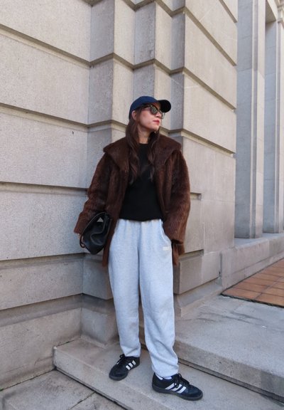 Mujer con abrigo de piel marrón, top negro, pantalones deportivos grises, zapatillas negras Adidas, gorra negra y gafas de sol, de pie junto a la esquina de un edificio de piedra.