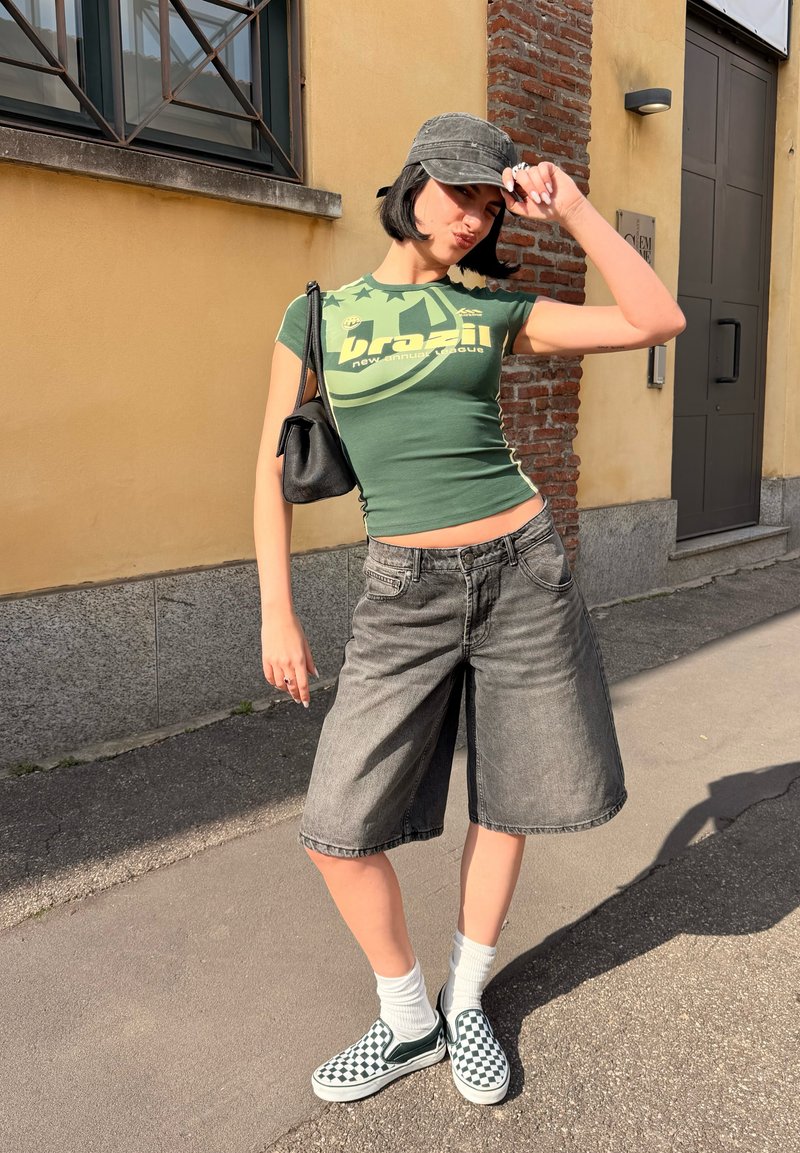 Jauns cilvēks zaļā T-kreklā ar uzrakstu "Brazil", pelēkos ceļos garos šortos, raibiem slip-on apaviem un melnu cepuri regulē cepuri ārā pie dzeltenas sienas.