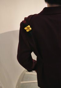 Veste bordeaux à texture lisse ; elle présente des accents en métal doré sur l'épaule. Escalier courbé visible en arrière-plan.