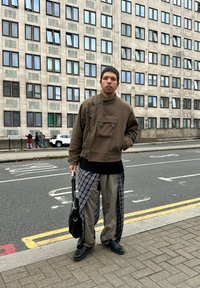 Vyras dėvi rudą striukę, languotus ir smėlio spalvos kelnes, juodus batus, laiko juodą krepšį, stovintis miesto šaligatvyje netoli kelio ir didelio pastato su langais.