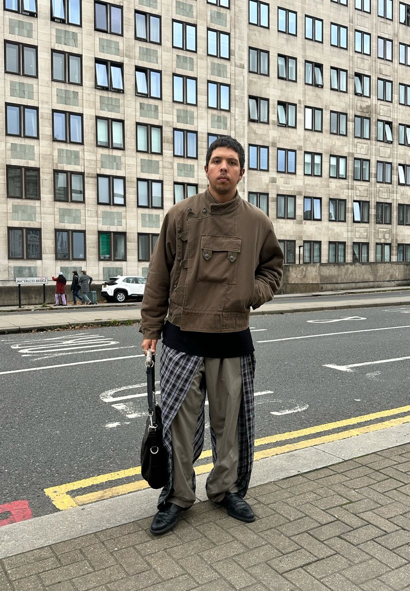 Man in brown jacket, plaid and beige pants, black shoes holding black bag, standing on city sidewalk near road and large building with windows.