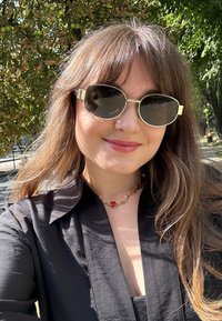 Στρογγυλά γυαλιά ηλίου με σκούρους φακούς και χρυσές λεπτομέρειες, συνδυασμένα με ένα μαύρο πουκάμισο με κουμπάκια και ένα κολιέ με κόκκινο κρεμαστό σχέδιο καρδιάς.