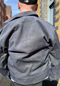 Σκούρο μπλε μπουφάν από χοντρό πανί με ριγωτή υφή, με κοντό γιακά και ορατές ραφές, απεικονιζόμενο από πίσω σε έναν τοίχο από τούβλα.