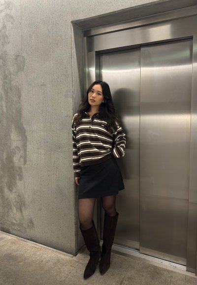 Mujer joven con un suéter a rayas, falda negra y botas apoyada contra una pared de concreto al lado de puertas de ascensor de acero inoxidable cerradas.