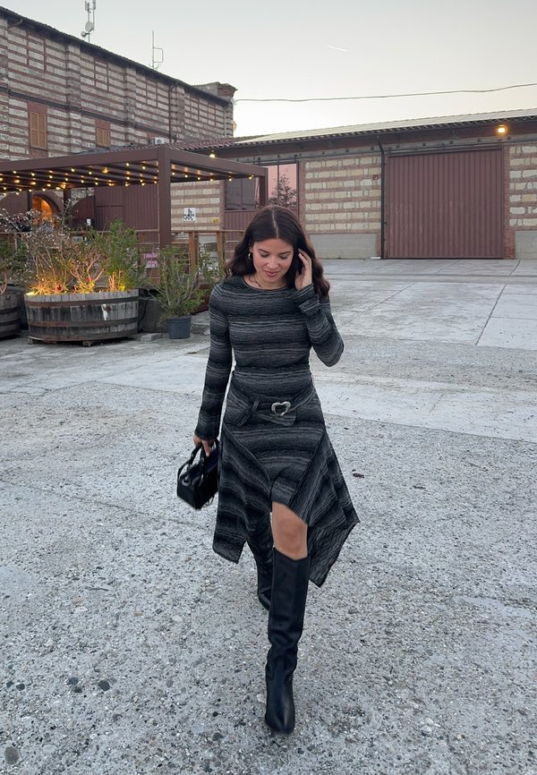 Mujer con vestido oscuro a rayas y botas negras caminando al aire libre cerca de edificios rústicos, sosteniendo un pequeño bolso negro y tocándose el cabello.
